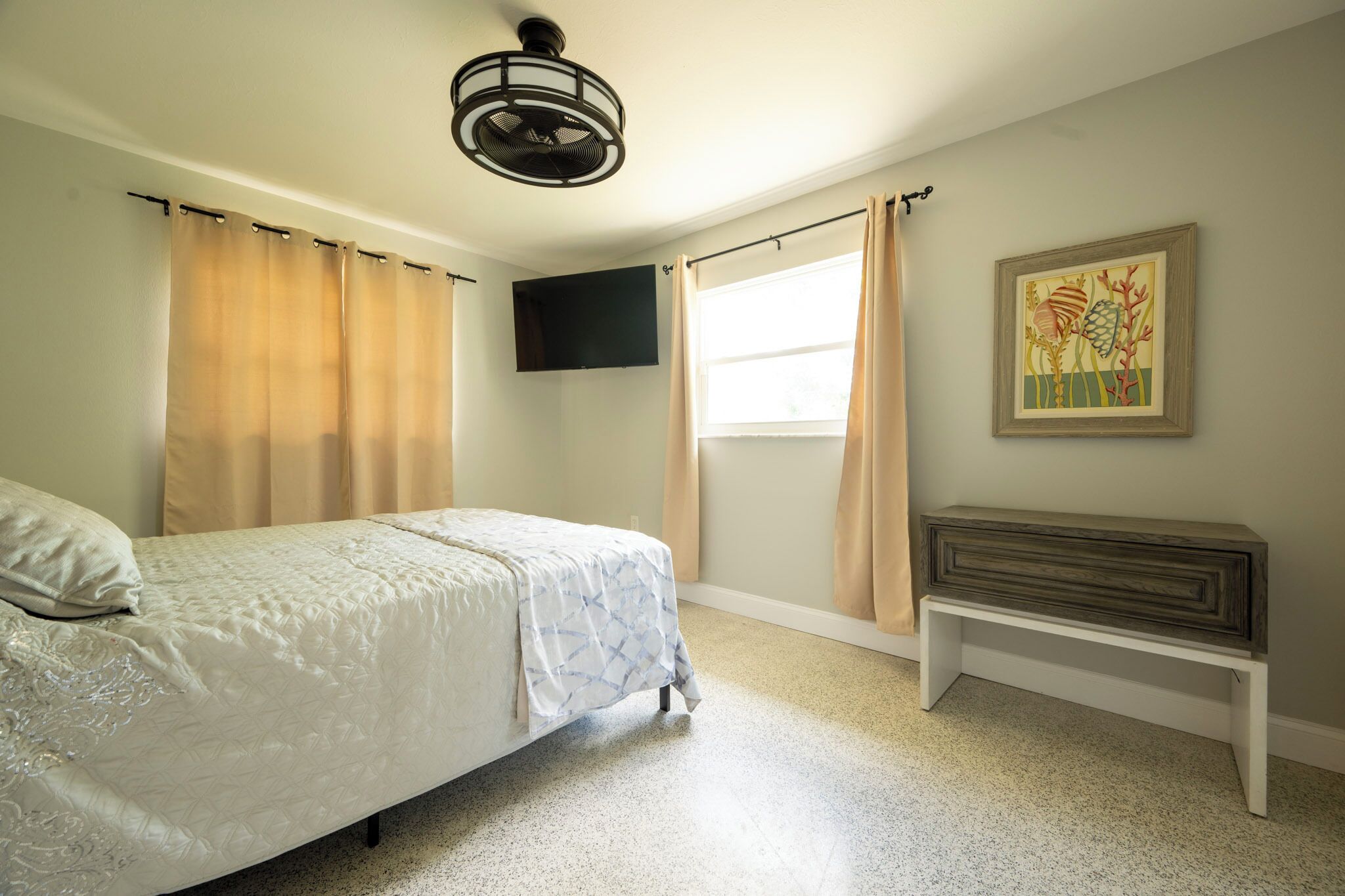 Un dormitorio con cama, ventilador de techo, ventana con cortinas, televisor montado en la pared y un banco.