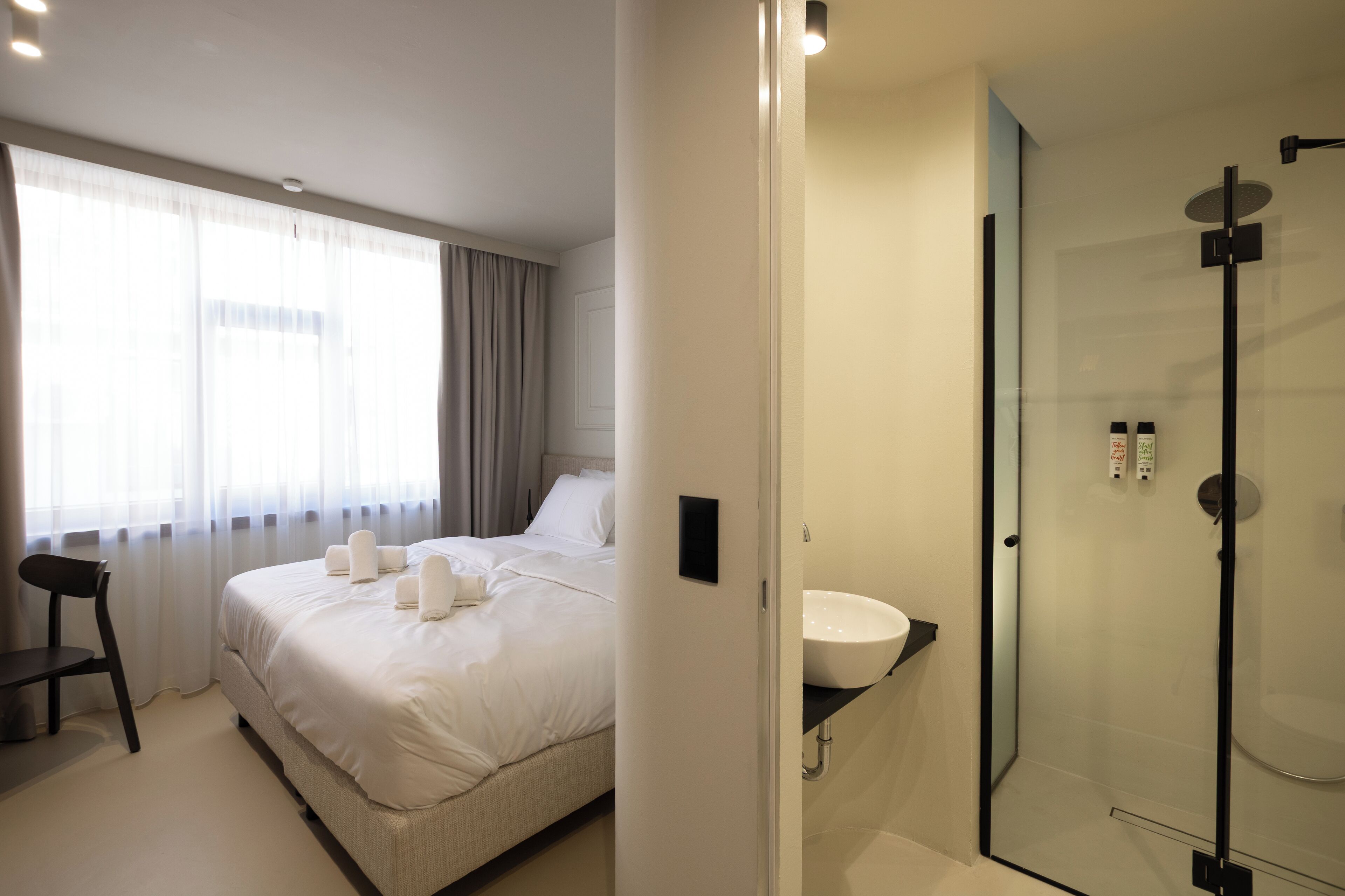 A hotel room with a bed, a chair, a bedside table, a window with curtains, and a glass-enclosed shower area.