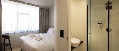 A hotel room with a bed, a chair, a bedside table, a window with curtains, and a glass-enclosed shower area.