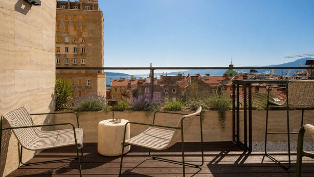 A balcony with wicker chairs, a small round table, and a view of buildings and a distant mountain. - Boutique Hotel OPERA (Rijeka)