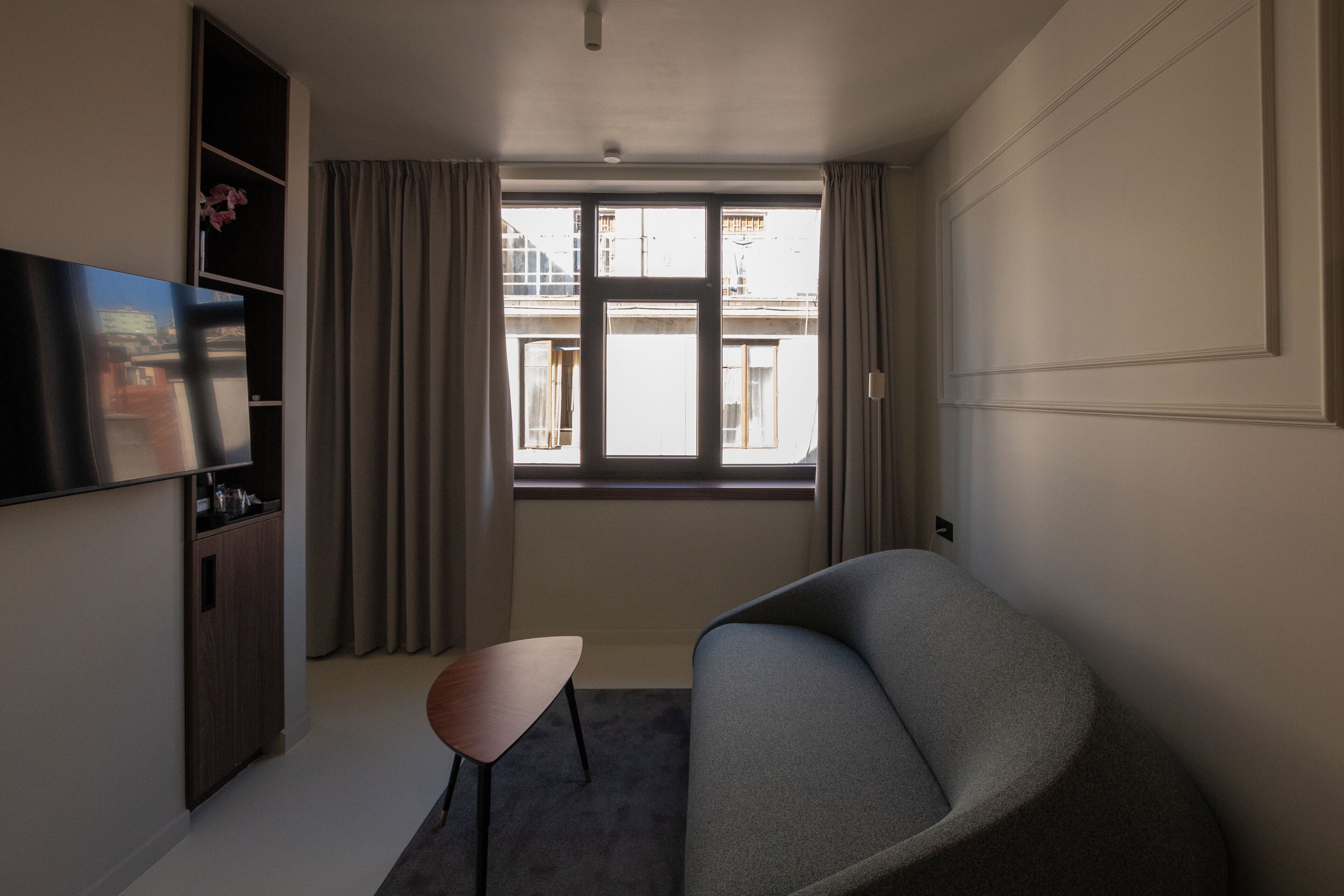 A modern living room with a grey sofa, a small wooden table, a flat-screen TV mounted on the wall, and a window with curtains.