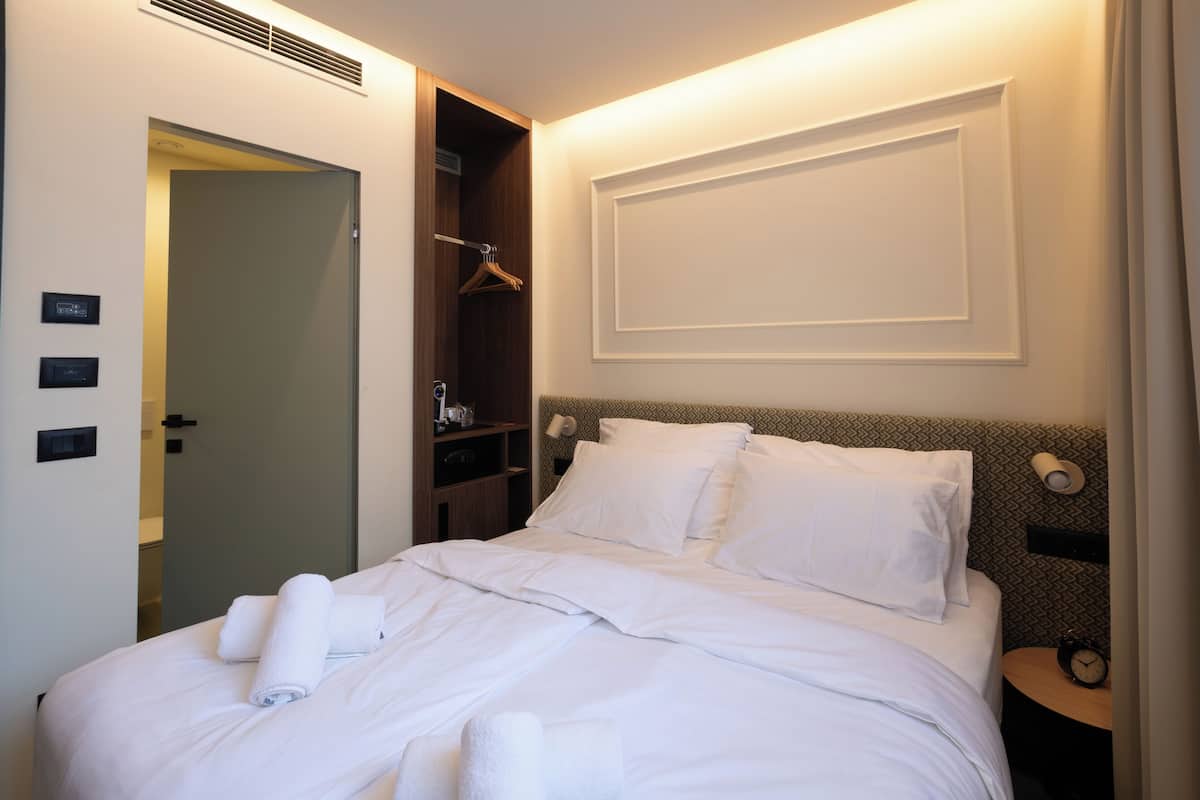 Un lit bien fait, avec du linge de lit blanc et des serviettes pliées, une armoire en bois et une table de chevet avec une lampe et un réveil.
