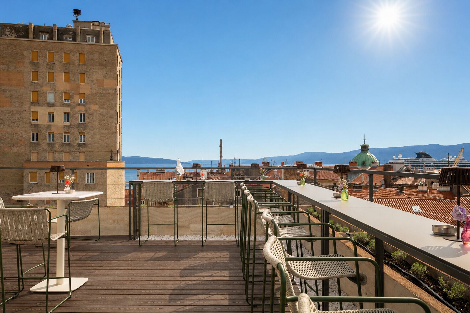 Eine Dachterrasse mit Außenmöbeln, Blick auf das Meer und ein großes Gebäude im Hintergrund.