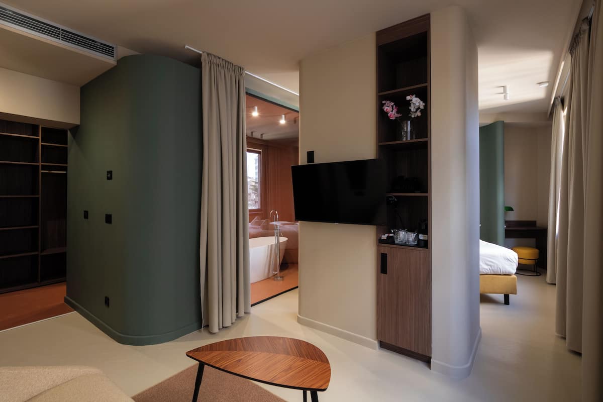Une chambre d’hôtel moderne avec un mur vert incurvé, un téléviseur à écran plat, une armoire en bois, un lit avec une tête de lit jaune et une salle de bain visible à travers une porte ouverte.