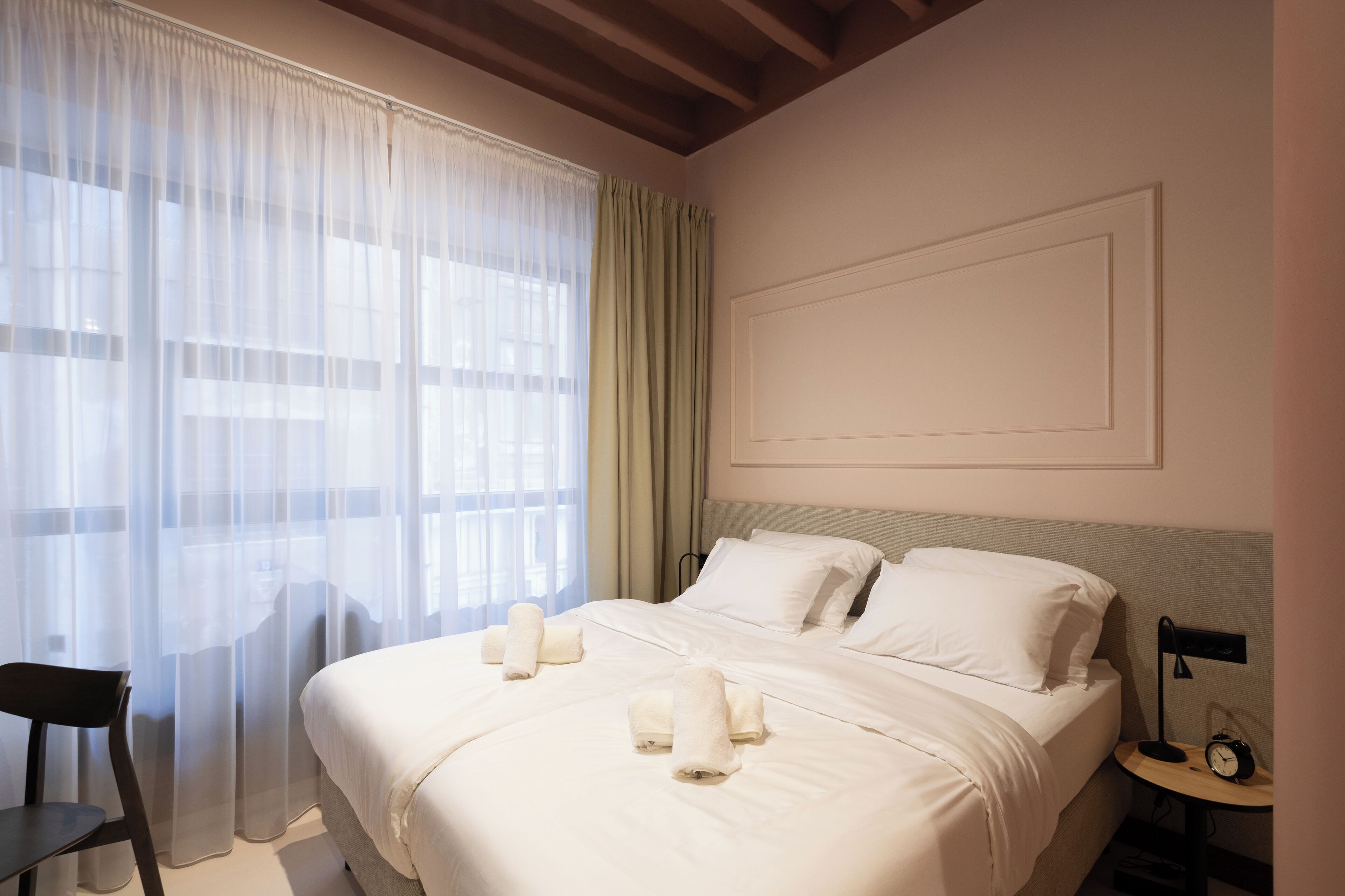Une chambre d’hôtel avec un lit, une chaise, une table de chevet avec une horloge et des rideaux légers.