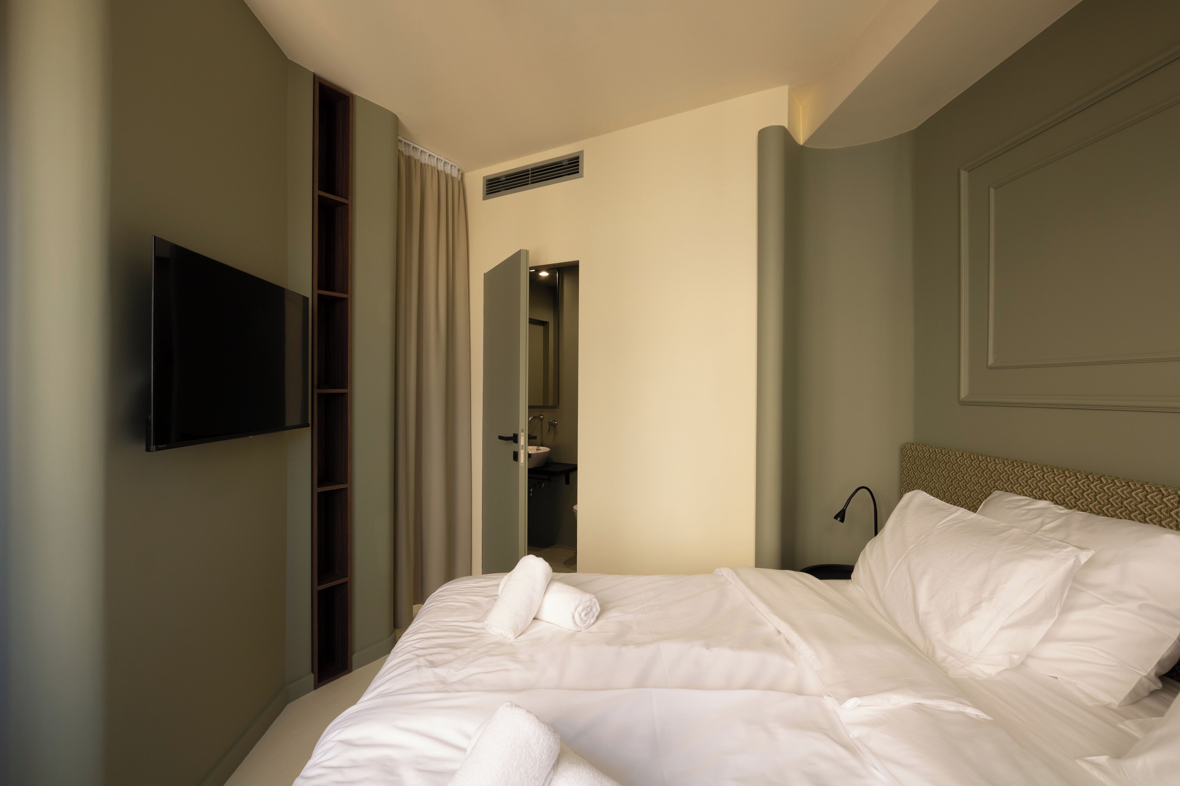Een hotelkamer met een bed, een aan de muur bevestigde televisie, een badkamer die door een open deur zichtbaar is en een ventilatie rooster.
