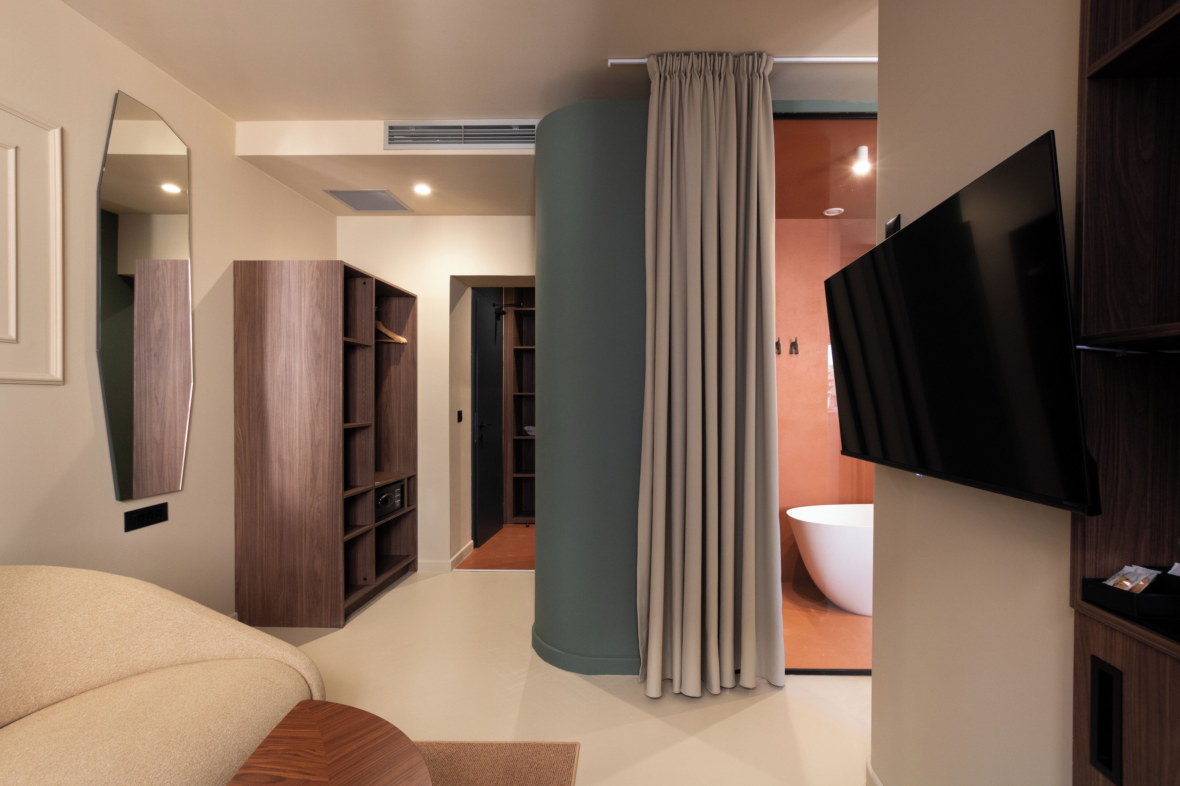 A modern bathroom with a bathtub, a toilet, and a shower area. There is a large mirror, a television mounted on the wall, and a wooden wardrobe.