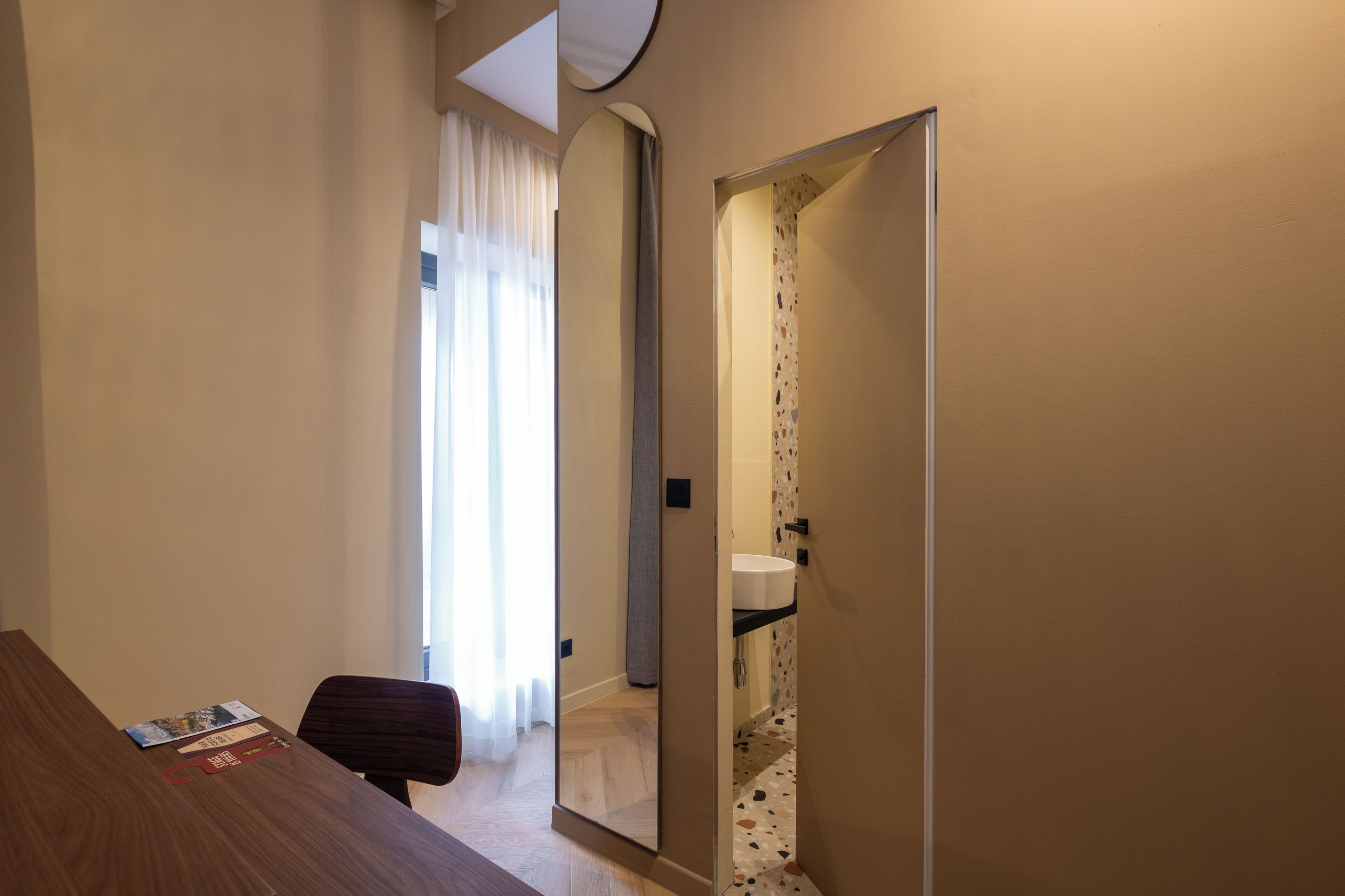 A hotel room with a wooden desk, a chair, a mirror, a bathroom with a sink, and a door leading to another room.