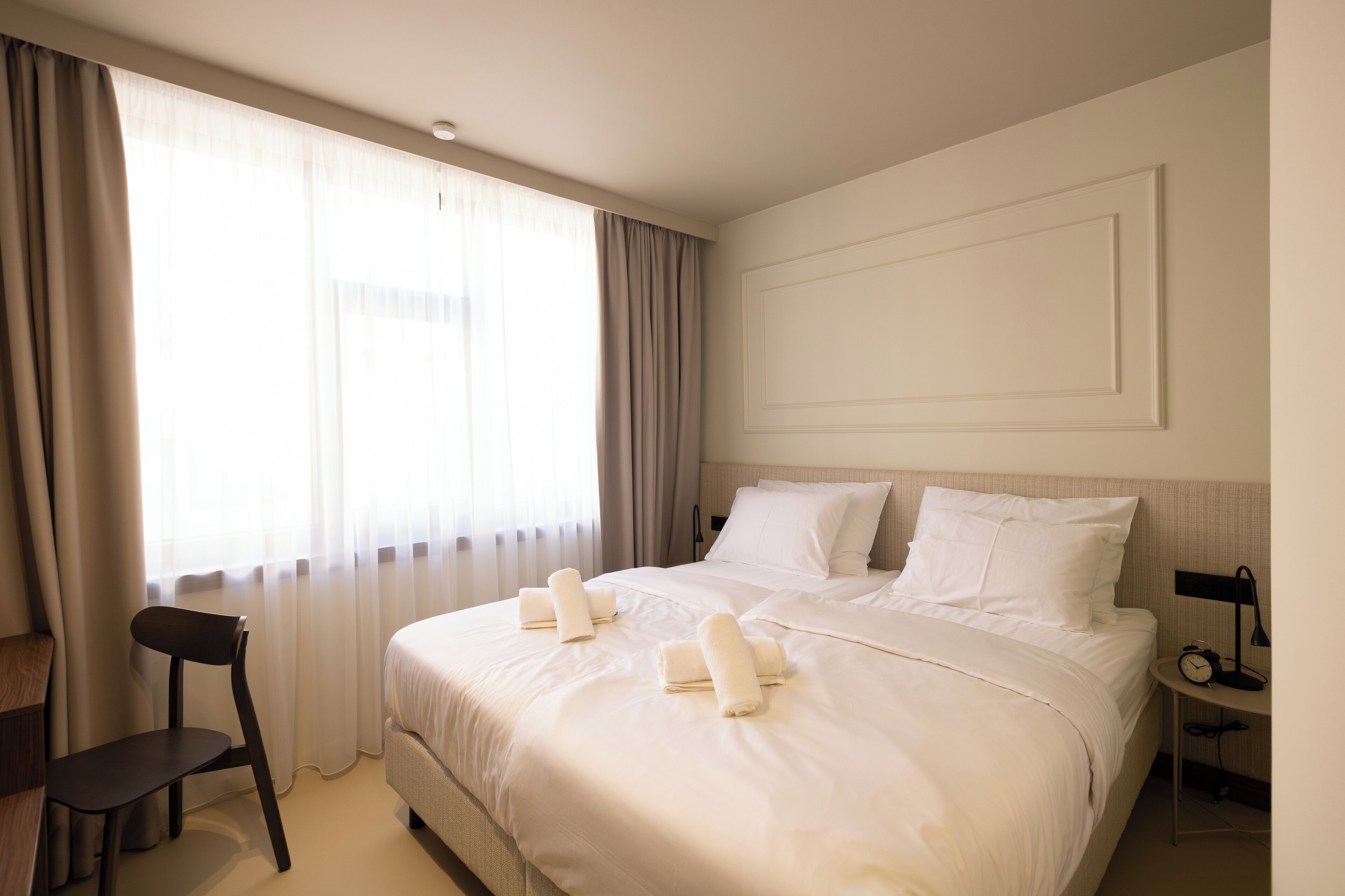 A hotel room with a bed, a desk, a chair, and a nightstand with a lamp.
