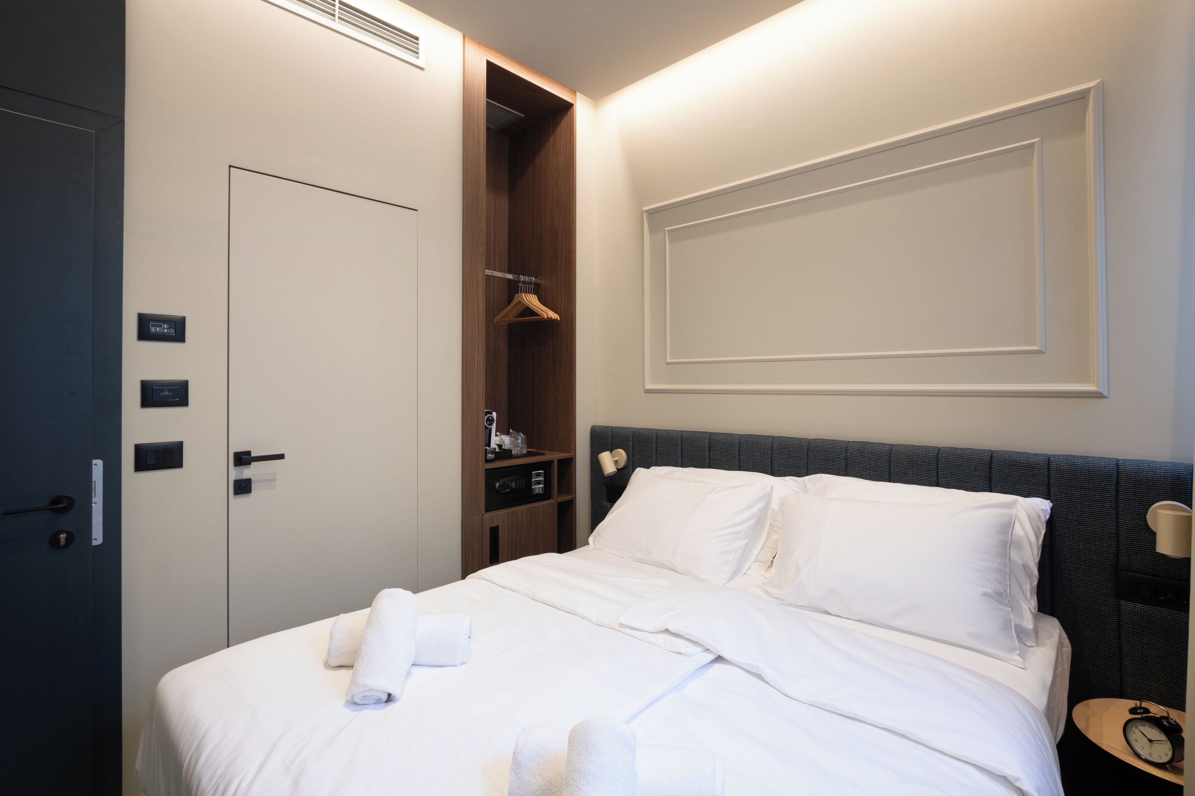 A hotel room with a bed, a wardrobe, a shelf with toiletries, and a bedside table with a lamp.