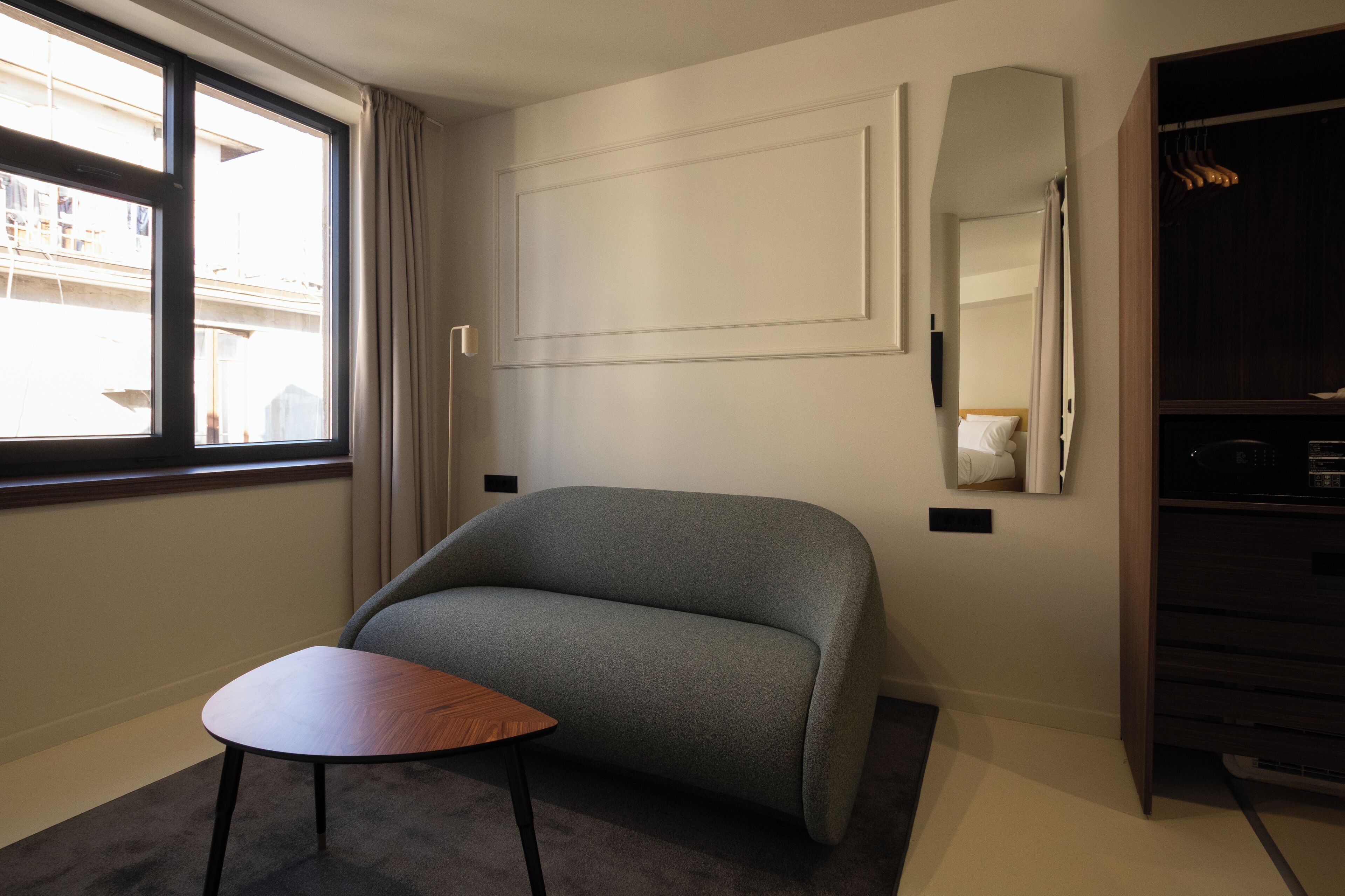 Une chambre d’hôtel moderne avec un canapé gris, une petite table en bois, une grande fenêtre avec des rideaux, une armoire intégrée et un miroir au mur.