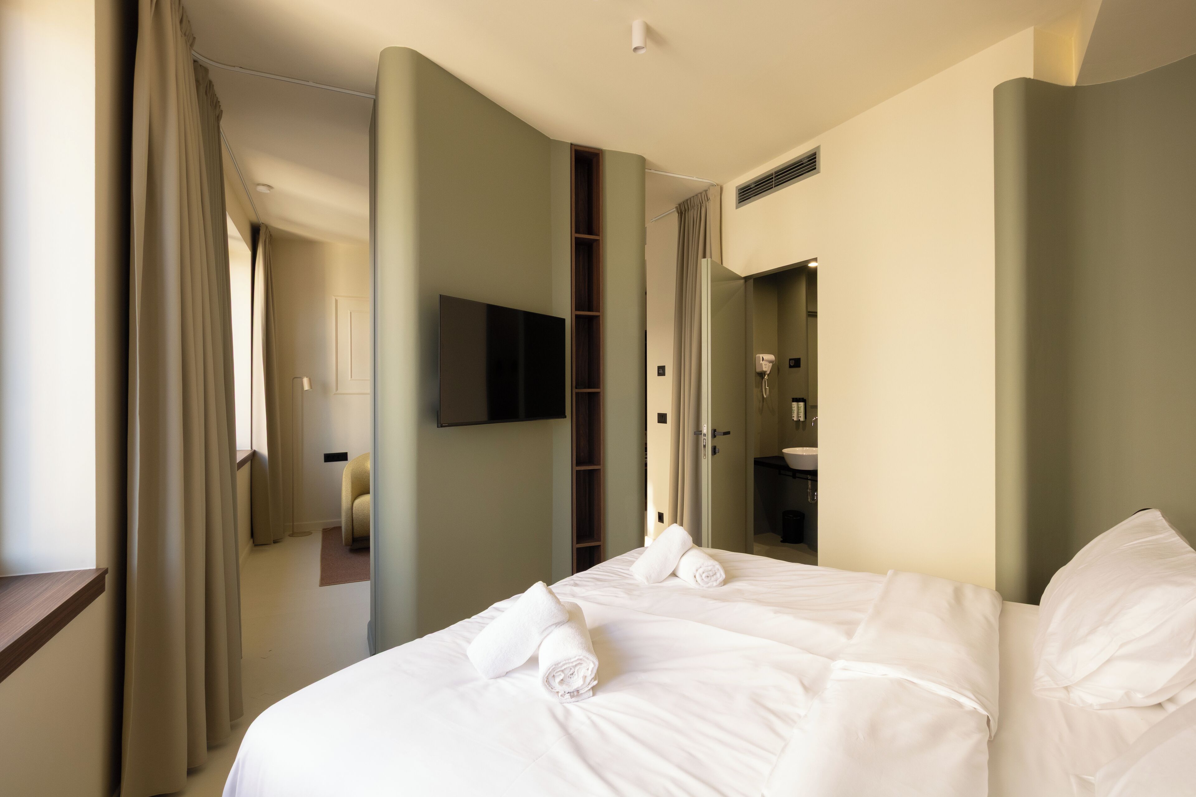 A hotel room with a large bed, a flat-screen TV mounted on the wall, a bathroom with a shower, and a small seating area.