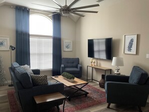 Un salon doté d’un ventilateur de plafond, d’une grande fenêtre avec des rideaux, d’un téléviseur à écran plat, d’une table basse et de deux canapés.