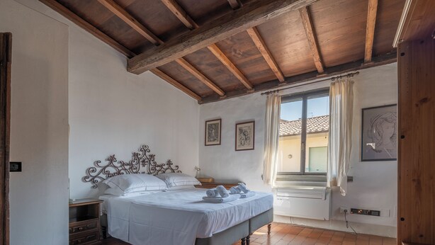 Ein Schlafzimmer mit Holzbalkendecke, einem Bett mit weißer Bettwäsche, einem Nachttisch und einem Fenster mit Vorhängen.