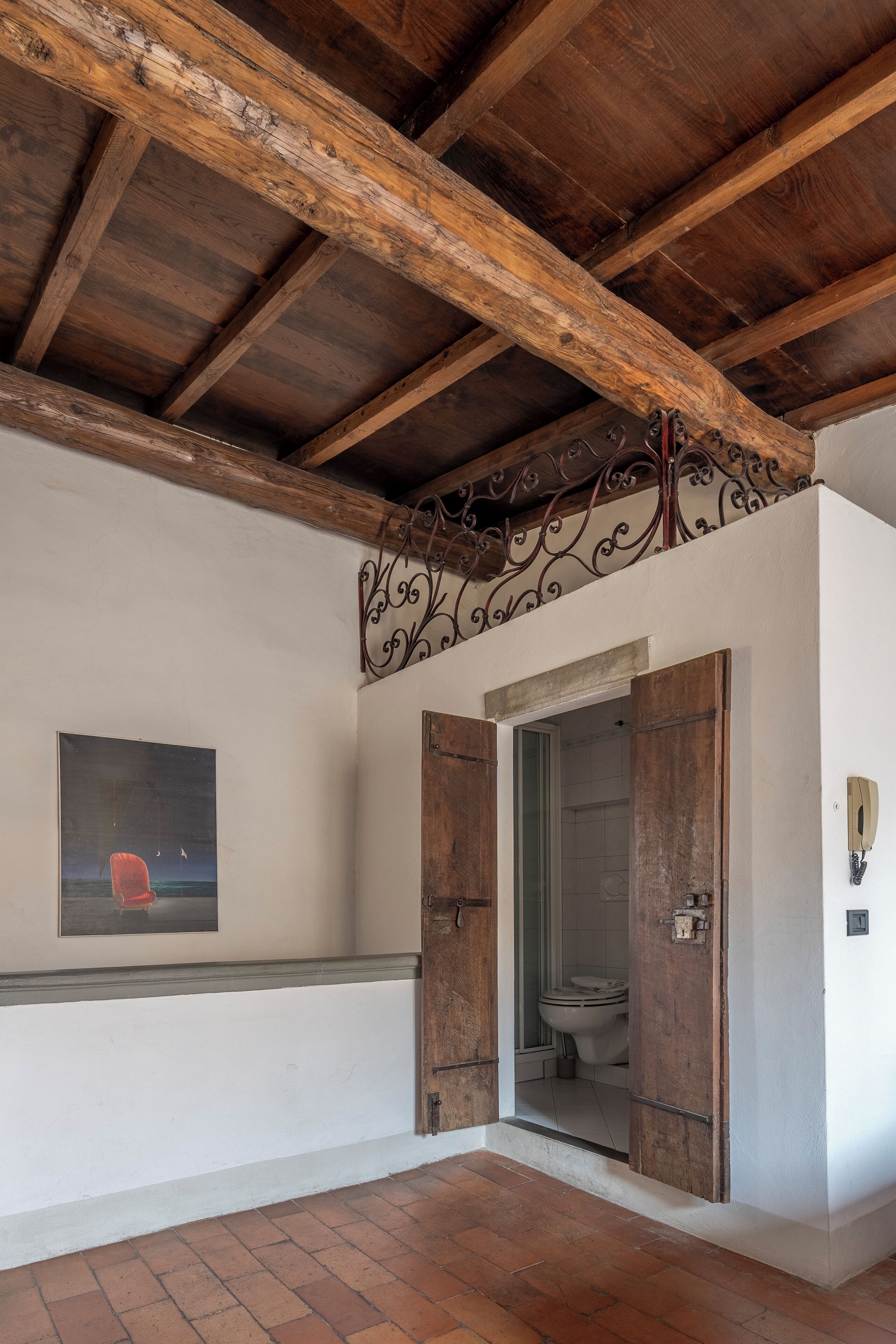 Een rustieke kamer met zichtbare houten balken, een witte muur met een ingelijst schilderij en een deur naar een badkamer.