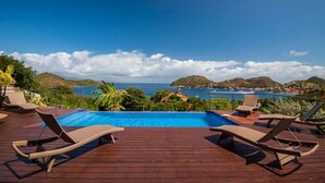 Een houten terras met lounge fauteuils, met uitzicht op een zwembad en de kust.