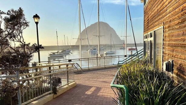 A waterfront walkway with a wooden building on the right, a clear view of boats and a mountain in the background. - Entire 2 Unit Duplex, King Beds, Rock View EV (Morro Bay)
