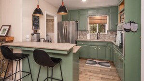 Uma cozinha com armários verdes, uma geladeira de aço inoxidável, um espaço de bar com banquetas, luminárias pendentes e um quadro negro com anotações.