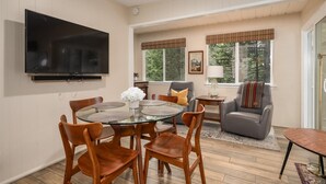 Uma sala de estar moderna com televisão de tela plana, uma mesa de jantar com cadeiras, um sofá cinza e uma janela com vista para árvores.