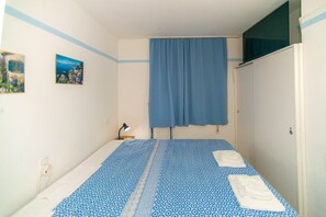 Una piccola camera con un letto singolo, copriletto azzurro fantasia, tenda blu e due quadri appesi al muro.