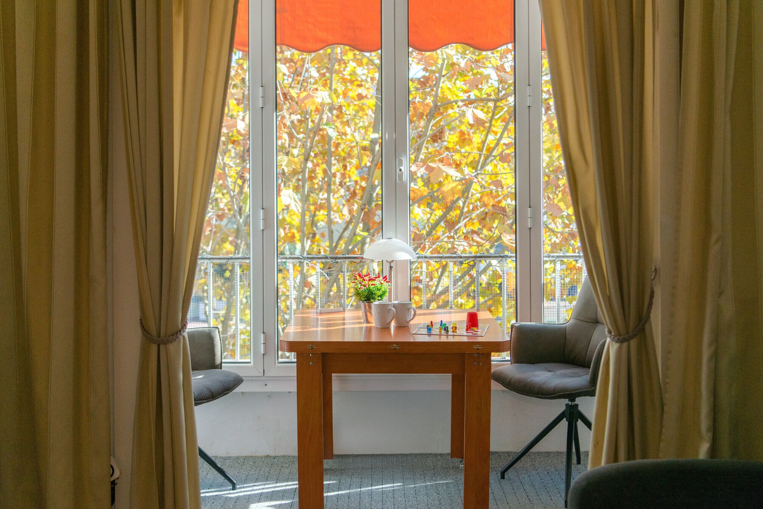 A room with a wooden table, two chairs, a potted plant, and a window with autumn leaves outside.