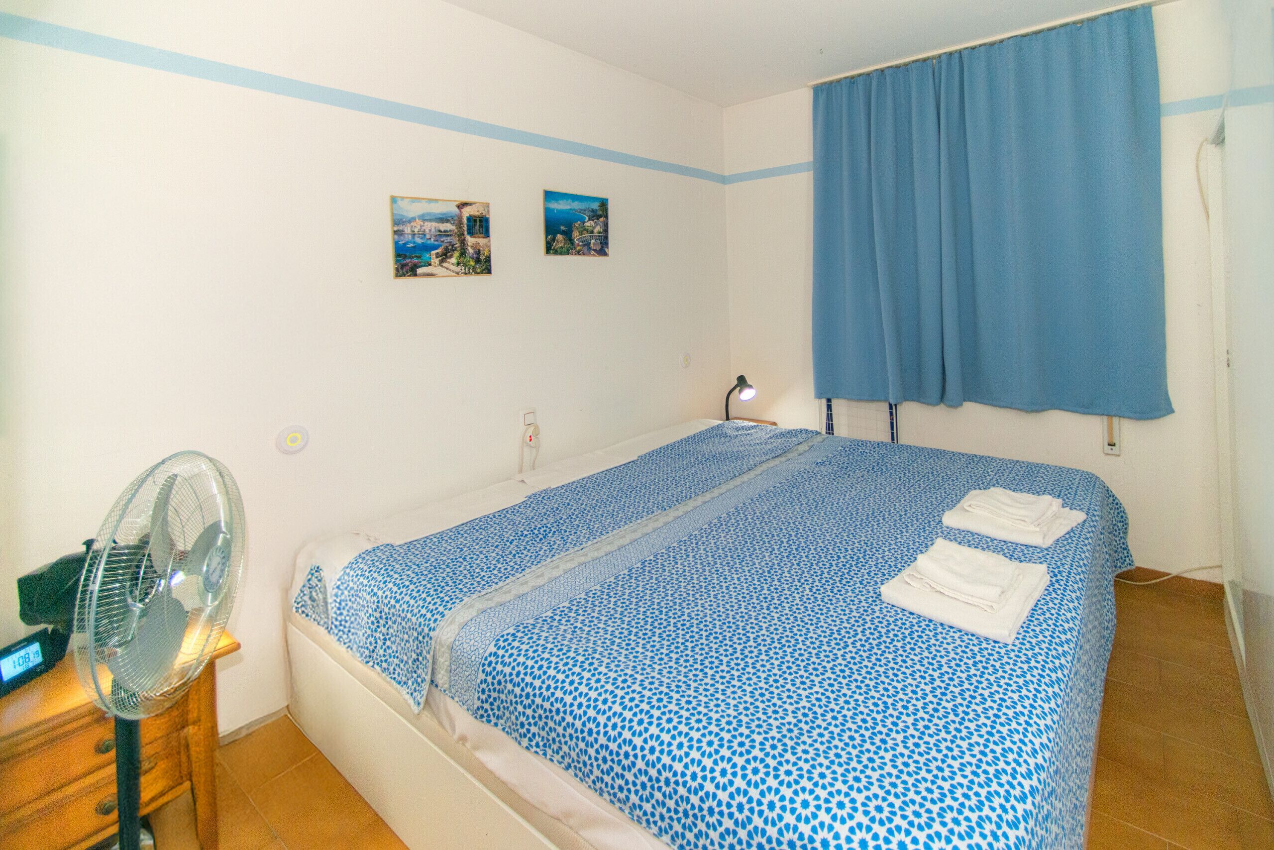 Ein Schlafzimmer mit einem Bett, einem Nachttisch, einem Ventilator und zwei Bildern an der Wand.