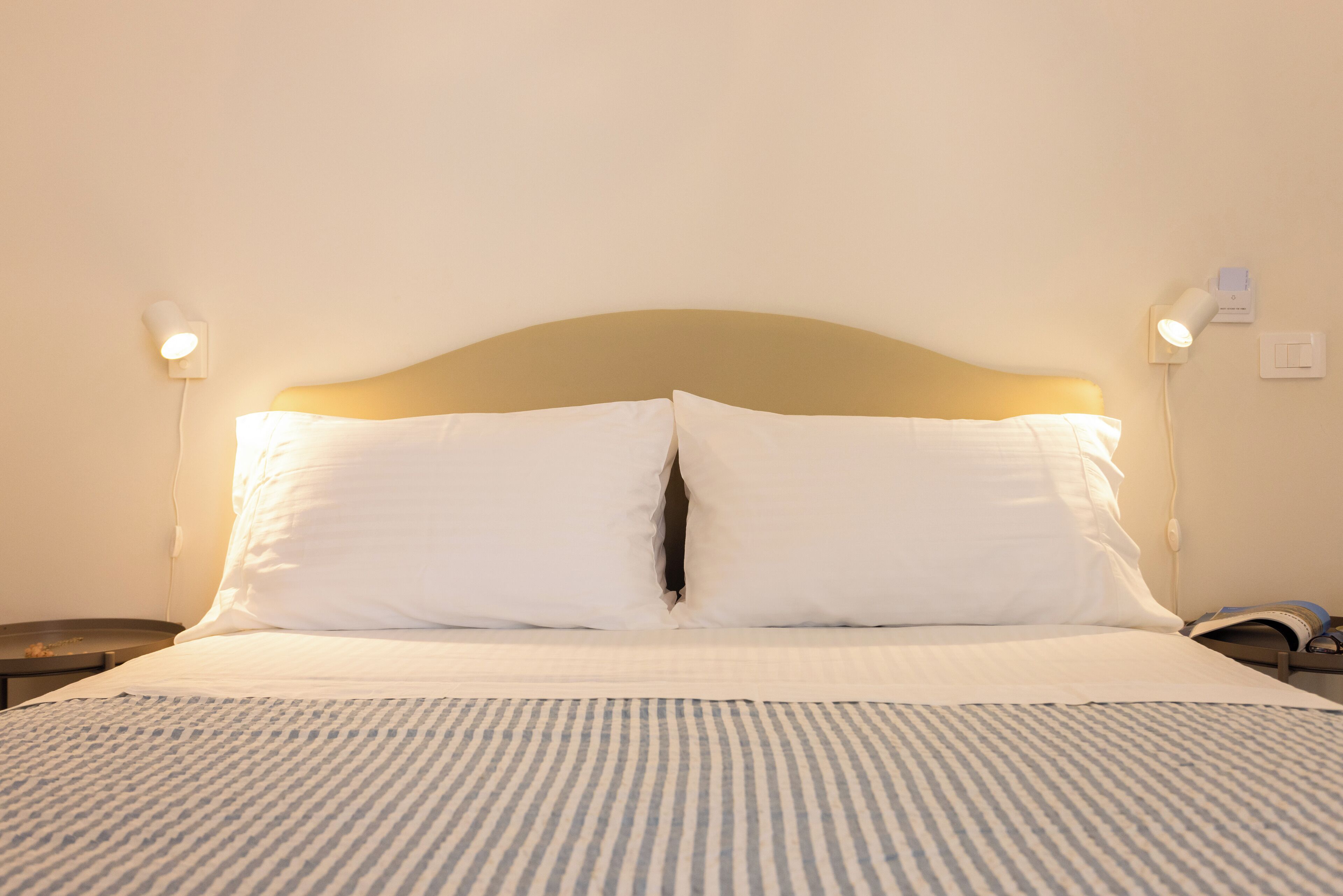 A neatly made bed with white linens and a striped bedspread, two bedside lamps, and a wall socket.