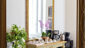 Ein Zimmer mit einem großen, verzierten Spiegel, einem kleinen Tisch mit Kaffeemaschine, einer Zimmerpflanze und einem Bücherregal.
