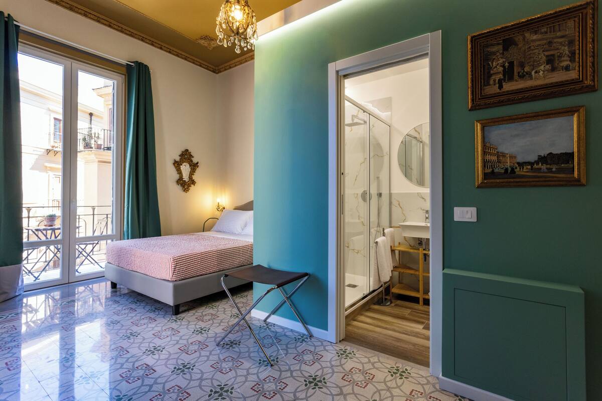 A bedroom with a bed, a small table, a chair, a bathroom with a sink and mirror, and a balcony with a view.
