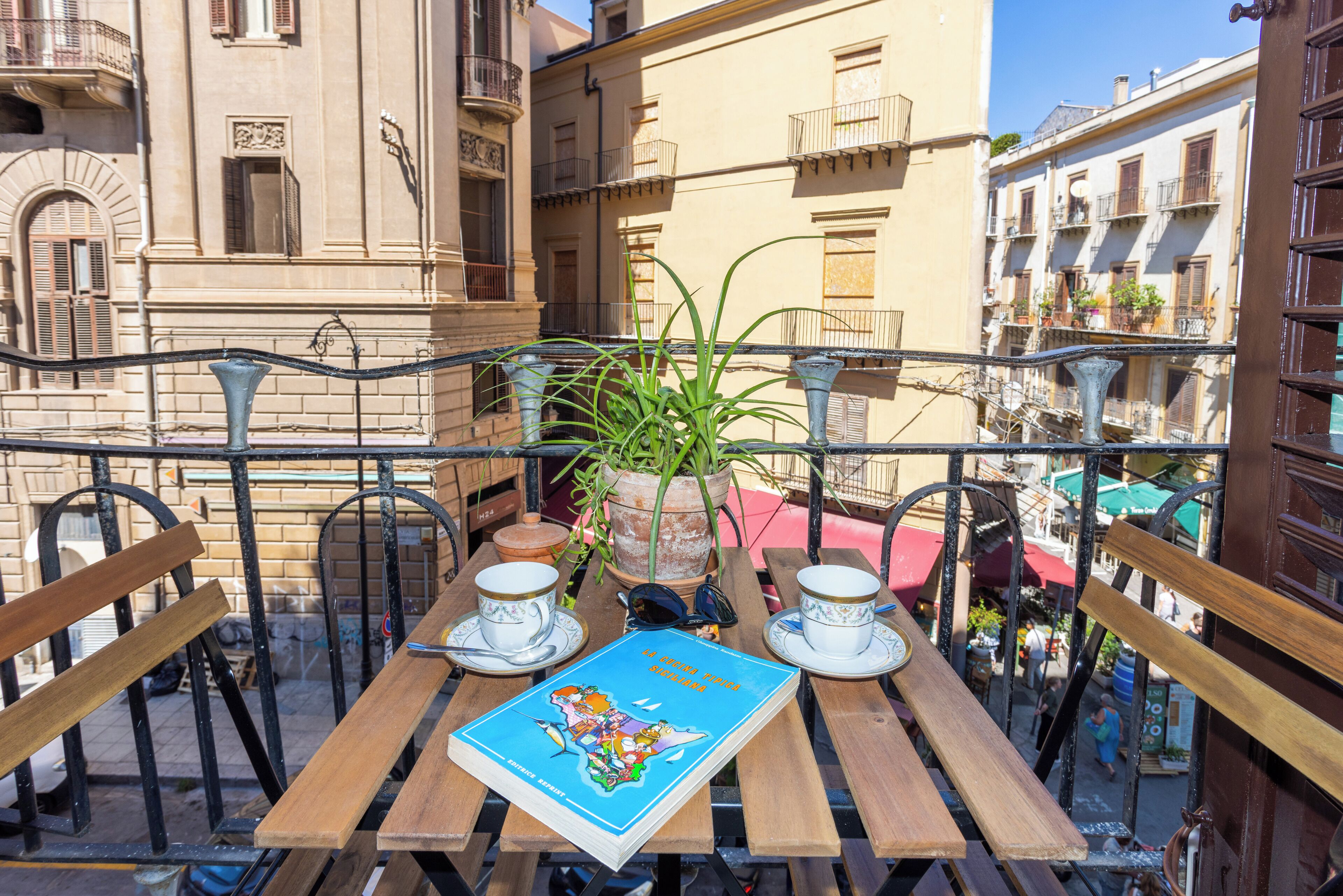 Una mesa de madera con un libro, dos tazas y una planta en maceta en un balcón con vistas a un paisaje urbano que incluye edificios y una calle.