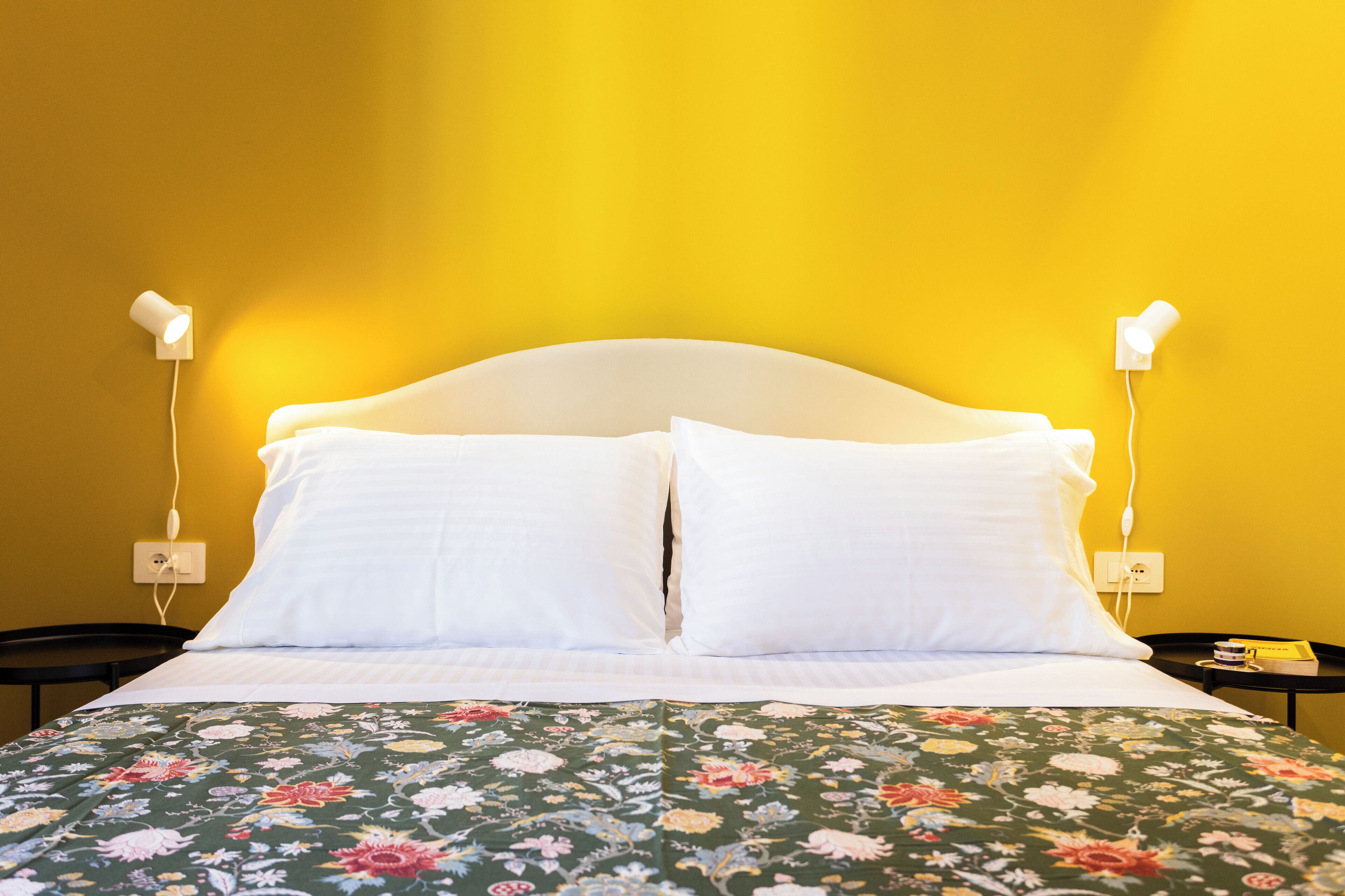 Cama com travesseiros brancos e colcha estampada, duas mesas de cabeceira com abajures e uma parede amarela.