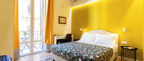 A hotel room with a bed, a desk, and a chair. There is a window with curtains, a wall-mounted air conditioner, and a small table with a lamp.