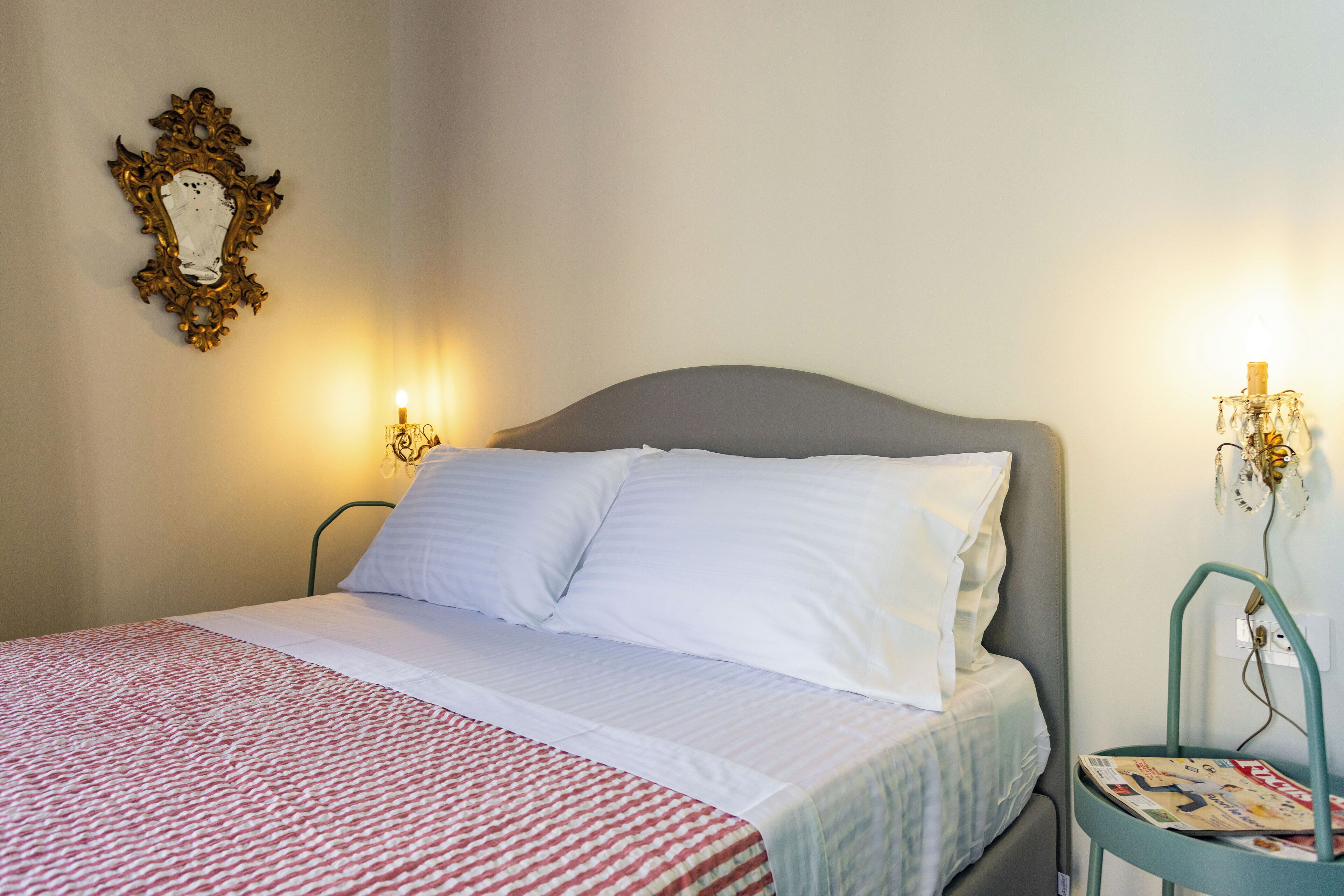 Una cama bien tendida con sábanas blancas, una manta a rayas rojas y blancas, y un espejo con marco dorado en la pared.