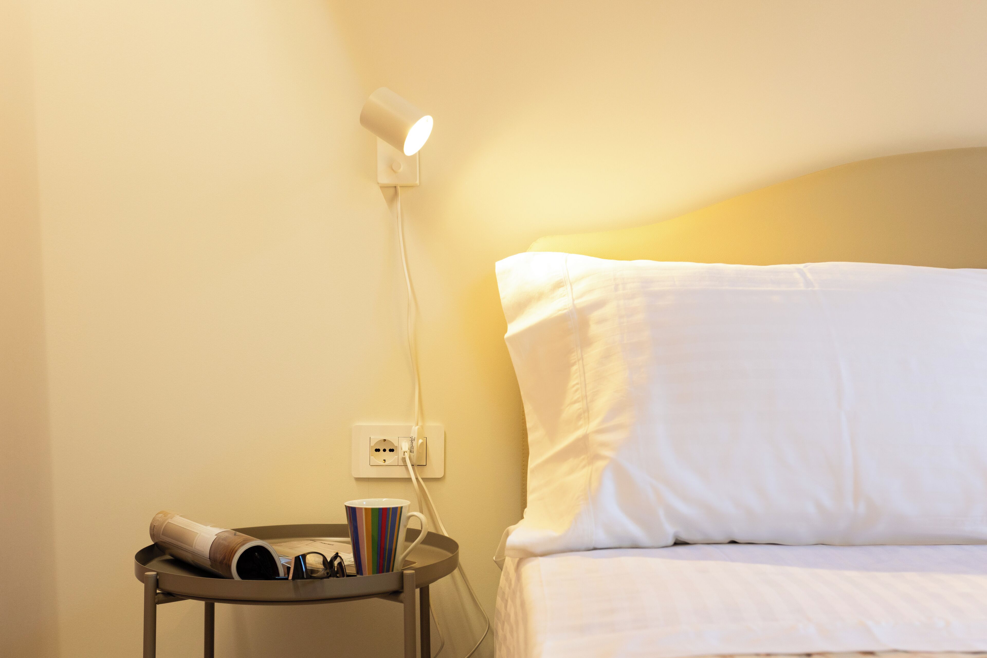 A neatly made bed with white linens, a bedside table with a cup and a magazine, and a wall-mounted lamp.