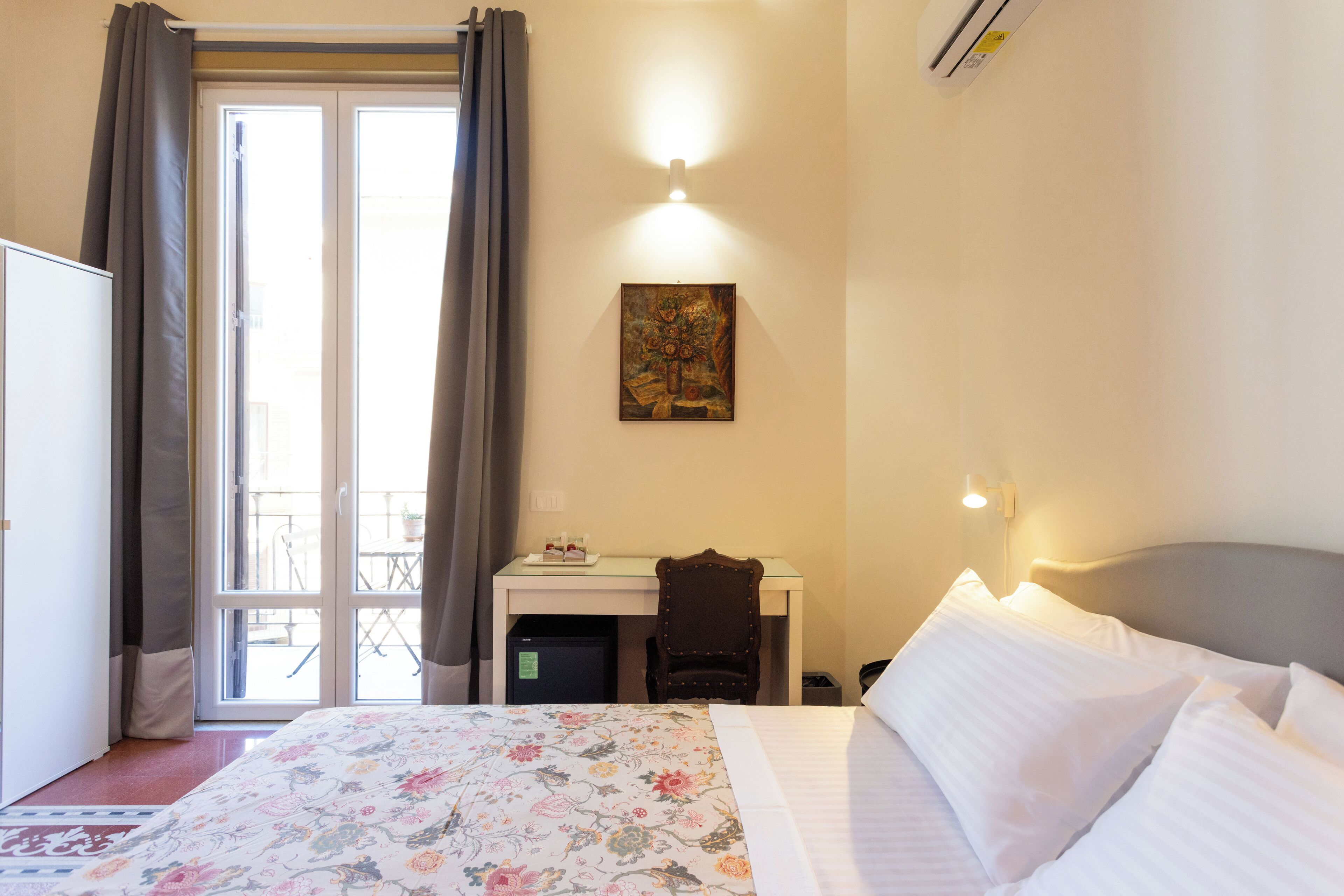 A hotel room with a bed, a desk, a chair, a small table, and a painting on the wall.