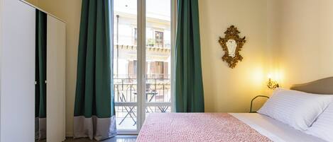 Una habitación de hotel con cama, balcón con cortinas verdes, lámpara de pared y suelo estampado.