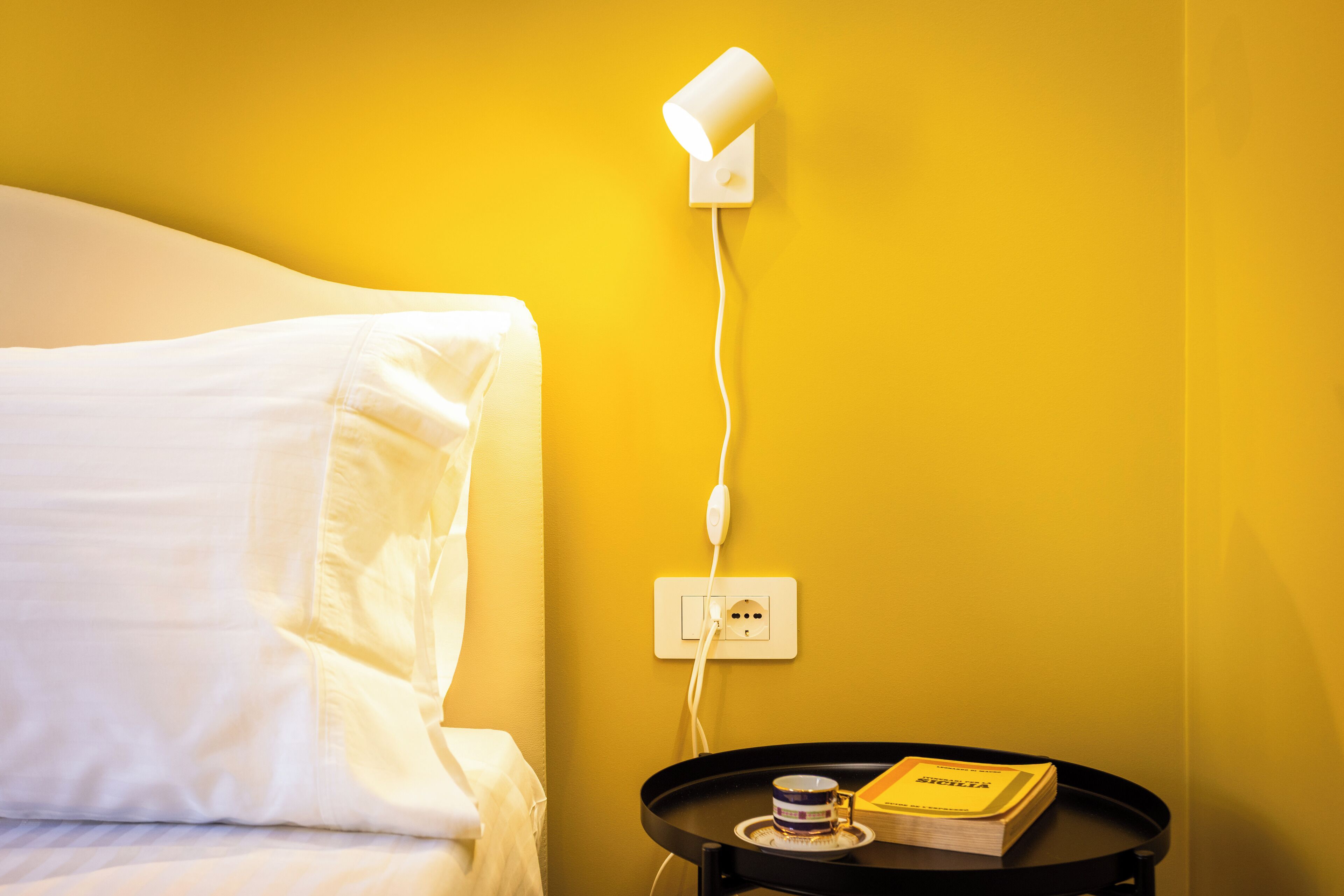A hotel room with a bed, a bedside table with a book and a cup, and a wall-mounted lamp.