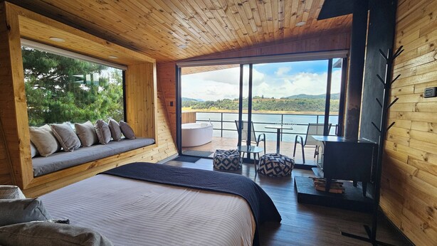 A cozy cabin with a large bed, a seating area with cushions, a fireplace, and a scenic view of a lake and hills. - NAQUA  (Chocontá)
