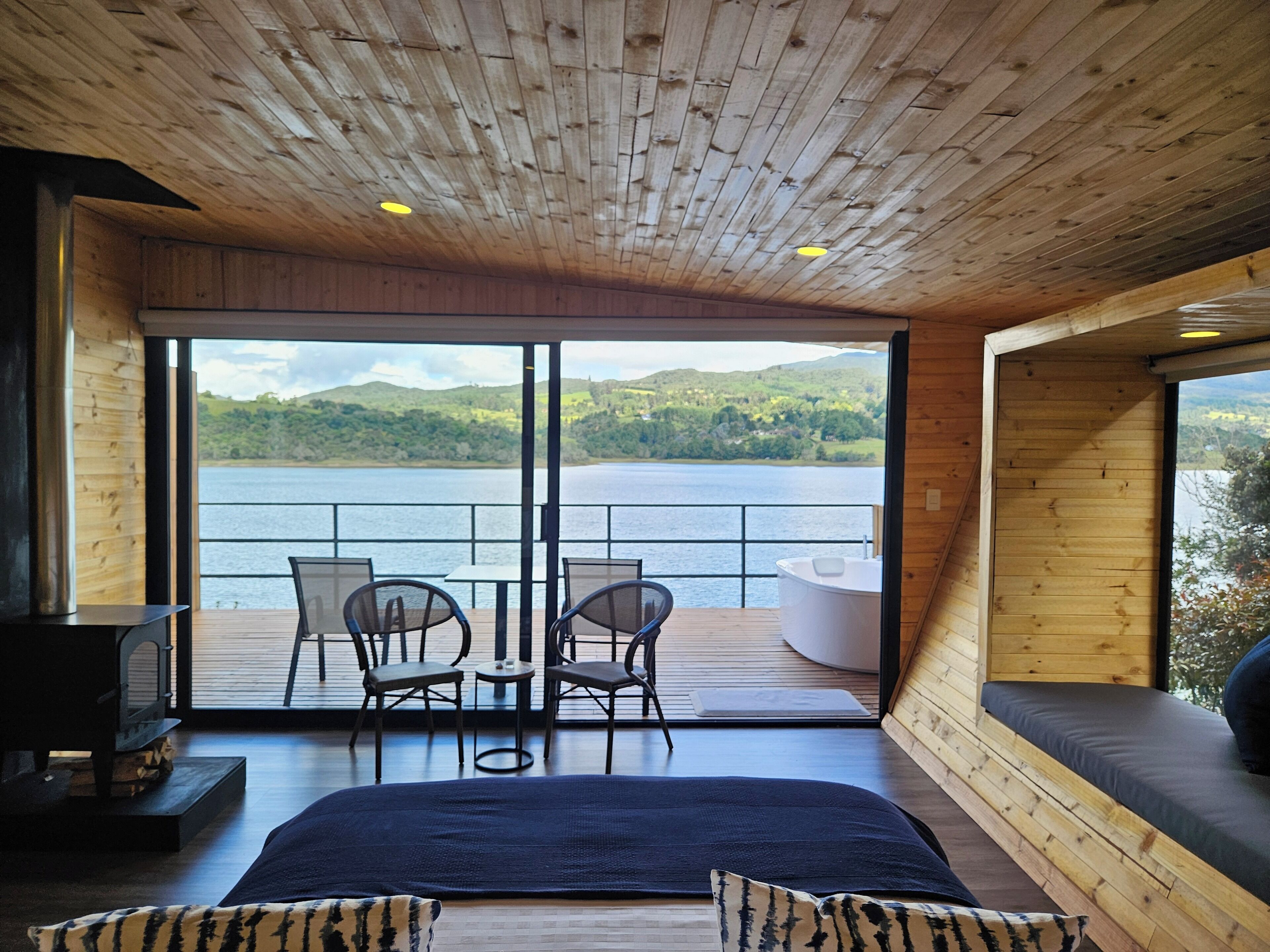 A wooden cabin with a large window overlooking a lake and hills. Inside, there is a bed with a dark blue bedspread, two chairs, and a small table.