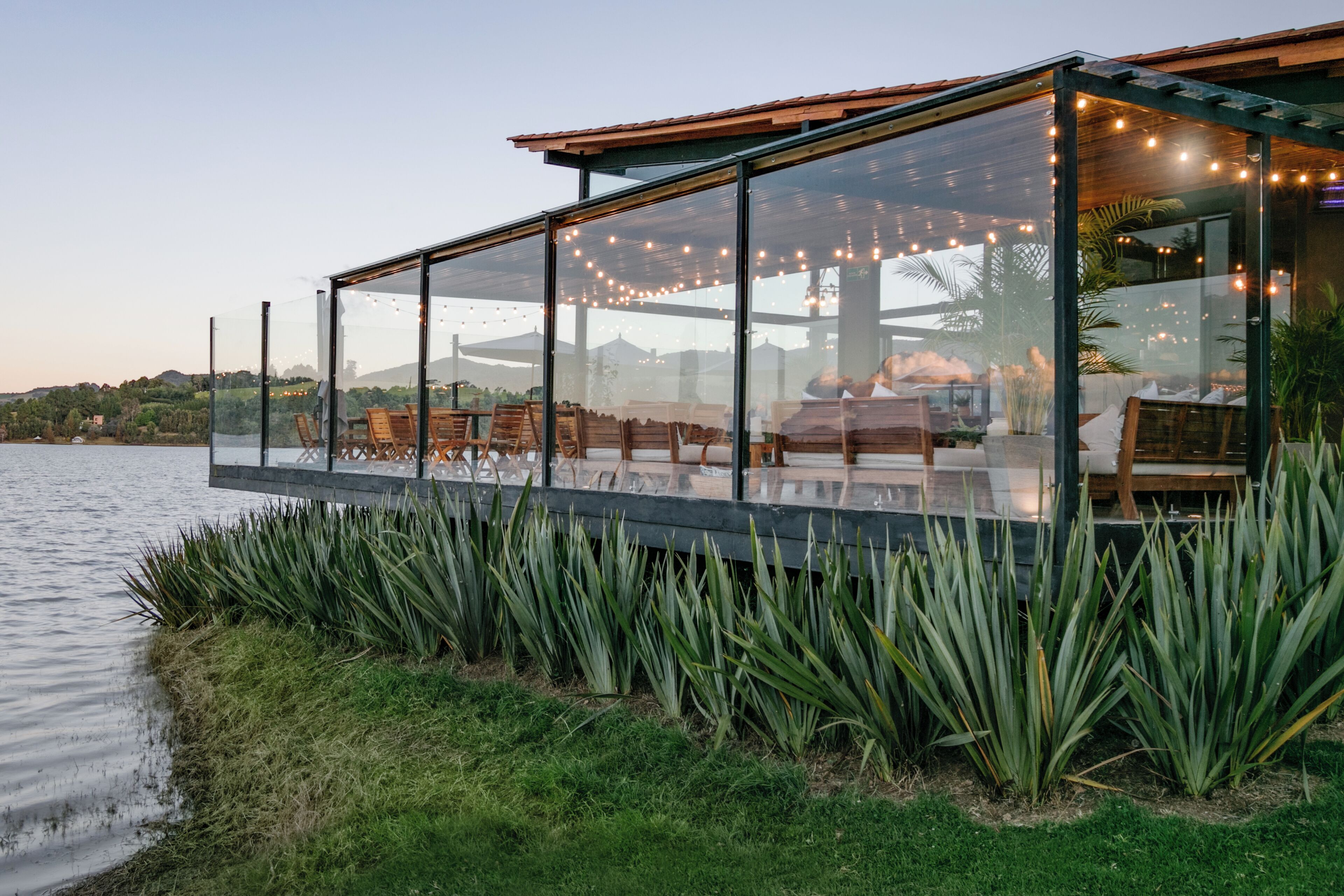 A glass-enclosed outdoor dining area with string lights, wooden furniture, and a view of the water and hills.