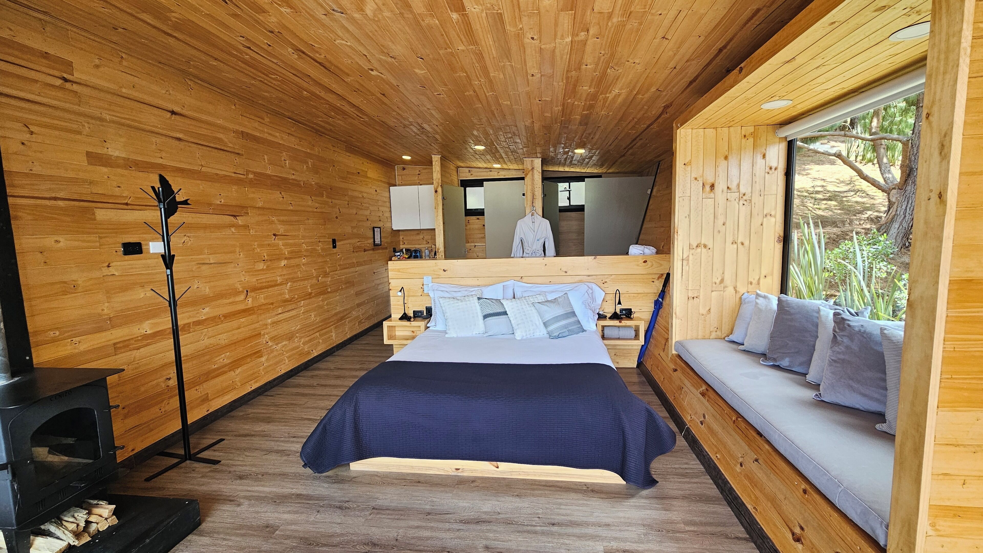 Interior de una cabaña de madera con cama, sofá, chimenea y ventana con vistas a la vegetación.