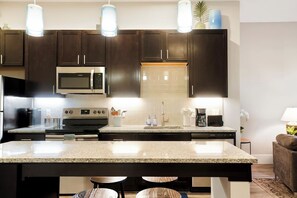 Une cuisine moderne avec des meubles de couleur sombre, un grand îlot central, des appareils électroménagers en acier inoxydable et des luminaires suspendus.