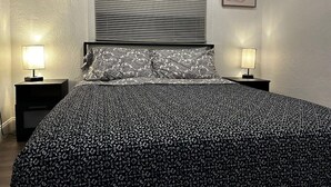 Een slaapkamer met een bed met een dessin, twee bedlampen, een raam met zonwering en abstracte wanddecoratie.