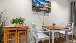 A dining area with a wooden table and white chairs, a mounted TV, and a potted plant.