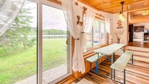 Un intérieur en bois avec une porte-fenêtre coulissante donnant sur une zone herbeuse, une table avec des bancs, et une cuisine visible à travers une porte ouverte.