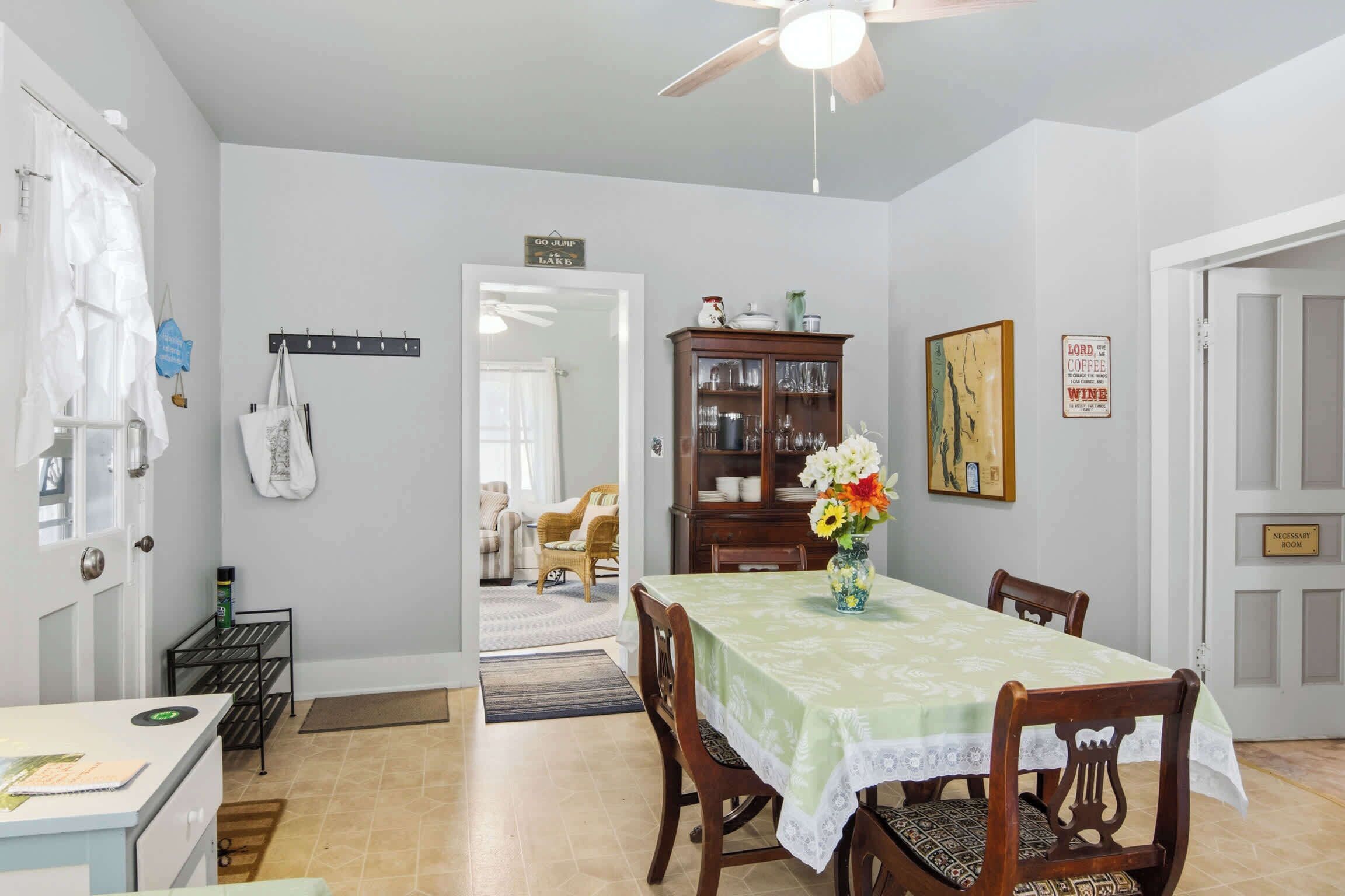 A dining area with a table set for four, a ceiling fan, a cabinet with glassware, and a door with a sign.