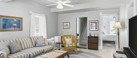 A living room with a striped sofa, a round coffee table, a wicker chair, a ceiling fan, and a television.