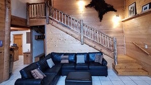 L’intérieur d’une cabane en rondins, avec un canapé d’angle noir, un escalier en bois et un ventilateur de plafond.