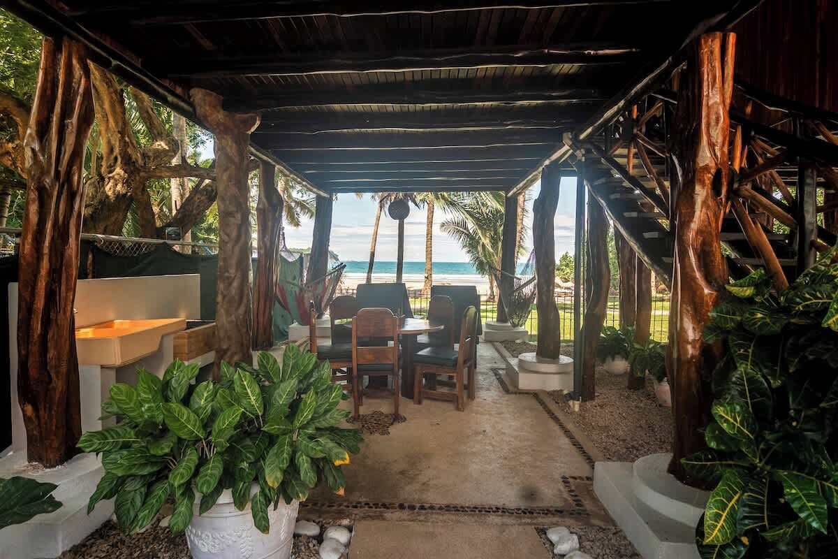 Un espace extérieur aménagé, abrité et doté de sièges, offrant une vue sur la plage et entouré de plantes tropicales et de structures en bois.