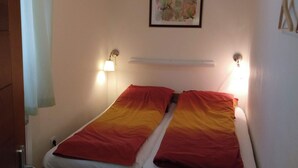 Ein Einzelbett mit einer roten und gelben Farbverlaufsdecke, zwei Wandleuchten und ein gerahmtes abstraktes Gemälde darüber.